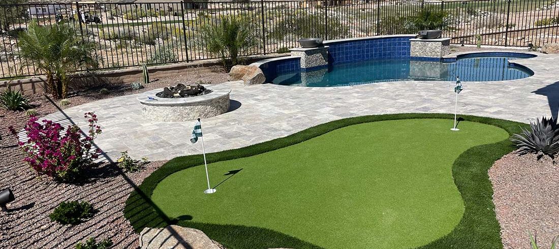 A backyard with a small putting green, a pool, a fire pit, and landscaped plants surrounded by a metal fence, with desert terrain and houses in the background.