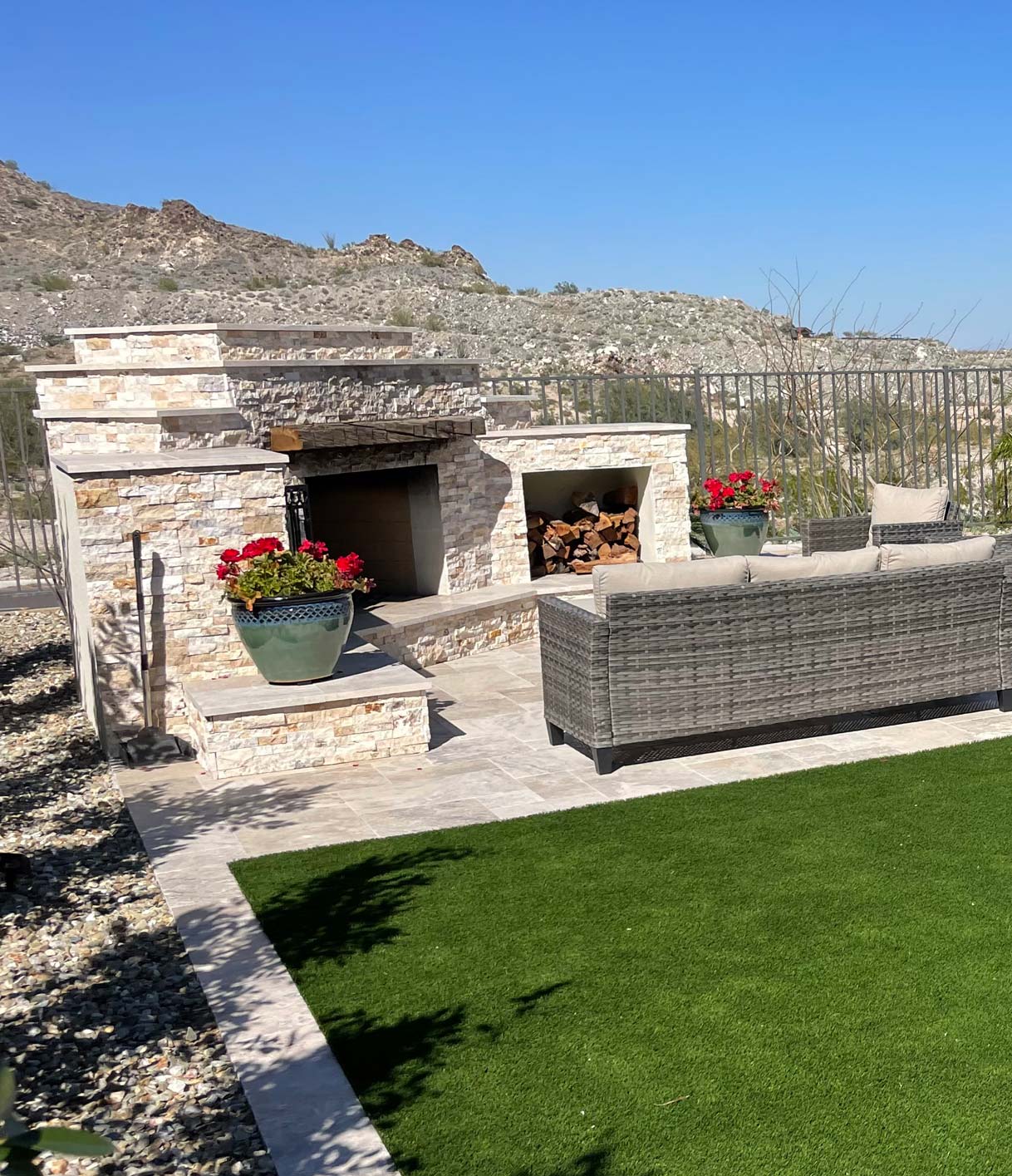 Outdoor patio with a stone fireplace, wicker furniture, and potted plants. The area overlooks a mountainous landscape under a clear blue sky.