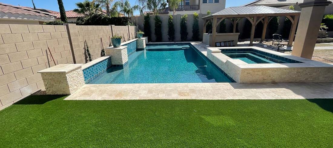 A backyard with a rectangular swimming pool, adjacent hot tub, and surrounding artificial turf, set in a residential area with a tiled-roof gazebo and palm trees.