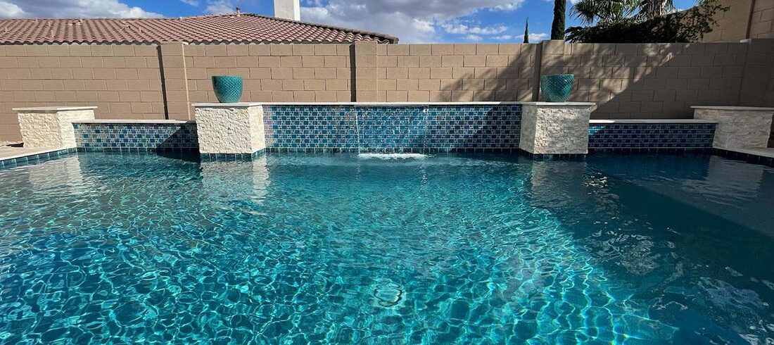 Backyard swimming pool with a stone tile water feature, two turquoise planters, and surrounding beige brick wall under a blue sky with scattered clouds.