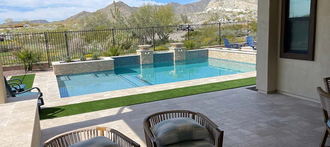 A backyard with a swimming pool, patio chairs, and a view of a desert landscape with mountains in the distance. The pool area is surrounded by a stone patio and a metal fence.