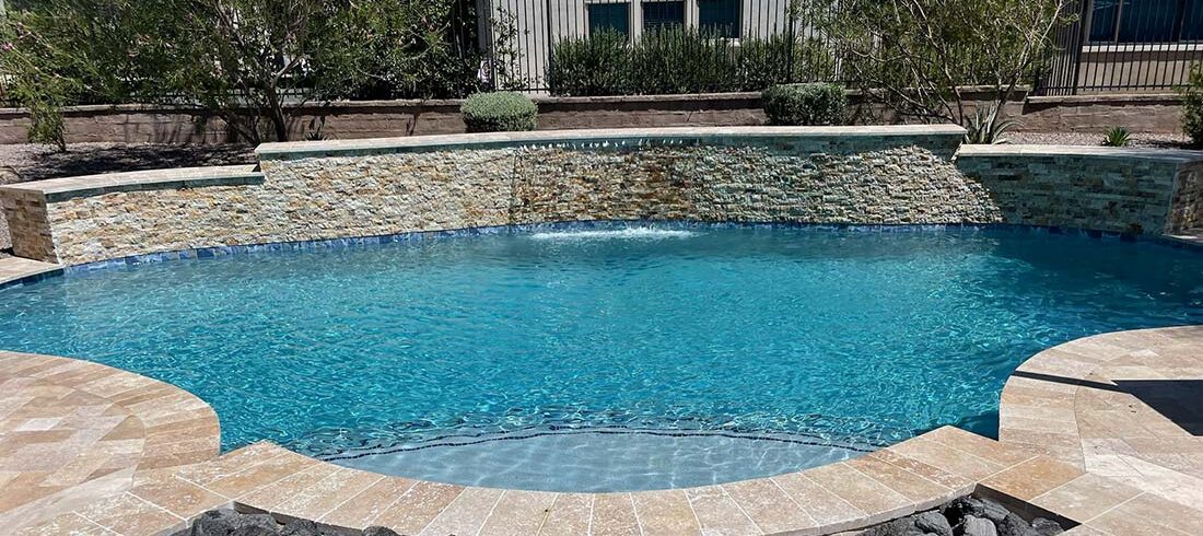 A clear blue swimming pool with a stone waterfall feature and tiled edges. Trees and greenery are in the background alongside a white building with windows.