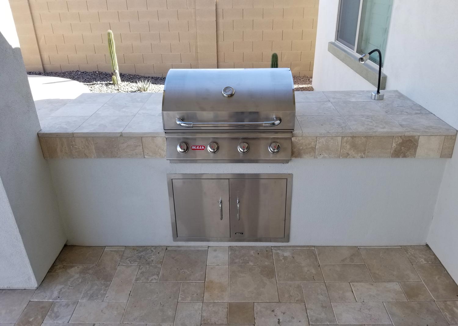 A built-in stainless steel gas grill with a tile countertop and a sink, set in an outdoor patio area with tiled flooring and a beige brick wall in the background.