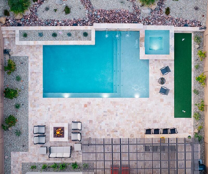 Aerial view of a backyard featuring a swimming pool, a hot tub, a fire pit with surrounding chairs, a small lawn area, and patio seating covered with a pergola.