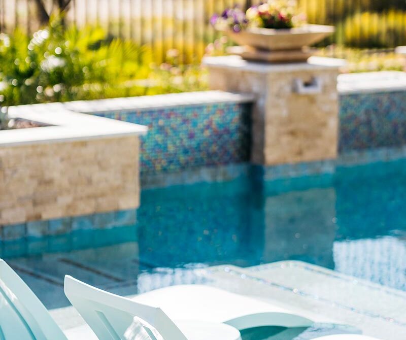 Two white lounge chairs are placed in a shallow section of a swimming pool, with a stone wall and a planter with colorful flowers in the background.