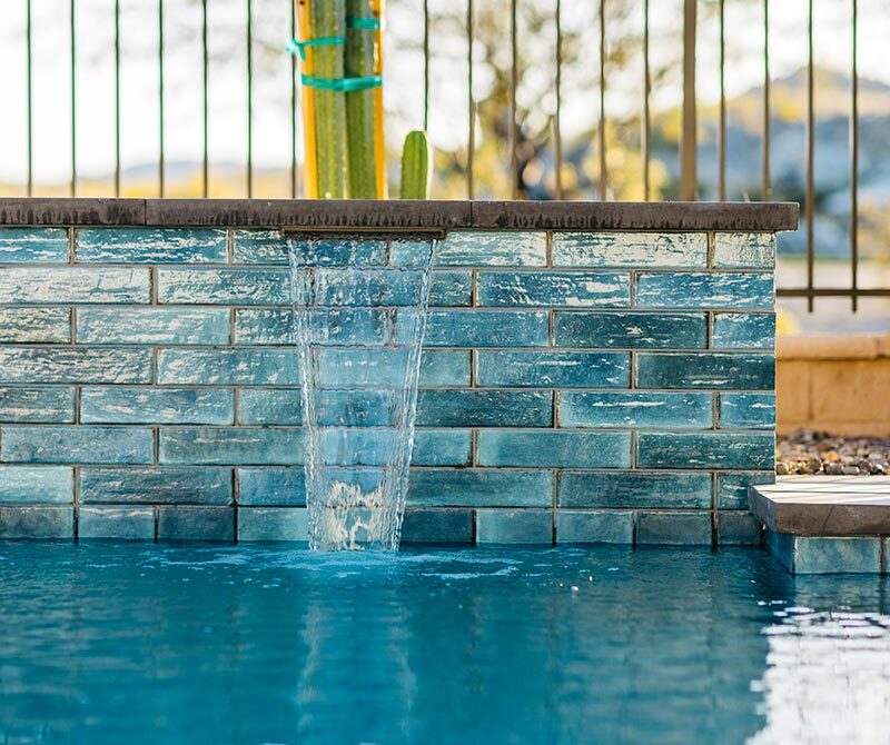 A swimming pool with a small waterfall feature, surrounded by a brick wall, and a cactus plant in the background near a metal fence.