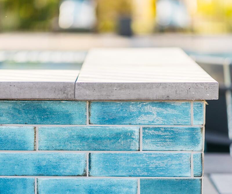 Side view of a pool edge with blue-green tiles and a concrete cap, set against a blurred outdoor background.