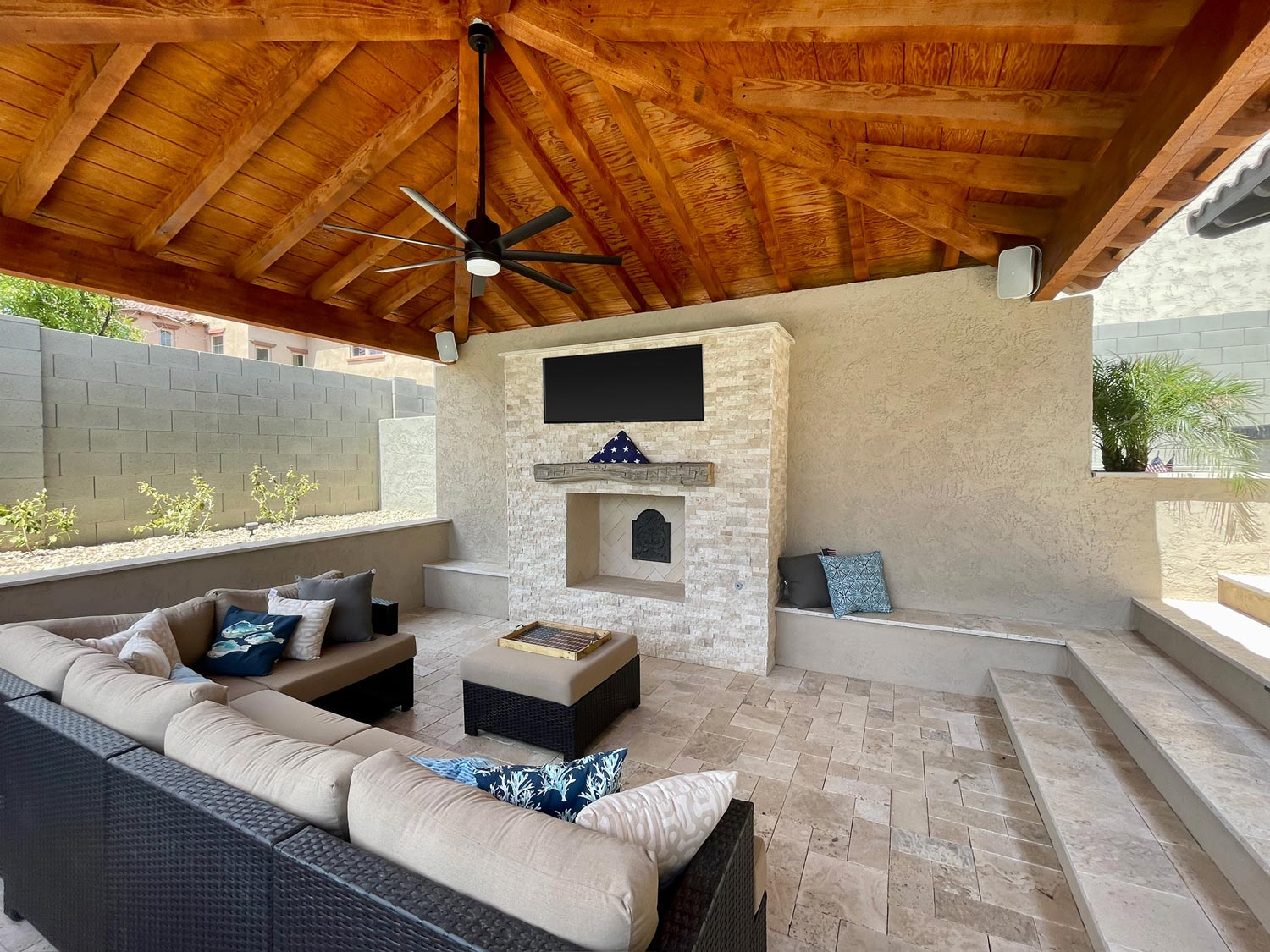 Outdoor patio with a wooden ceiling, a ceiling fan, a mounted TV above a stone fireplace, and a cushioned sectional sofa surrounding a coffee table with a small fire pit.