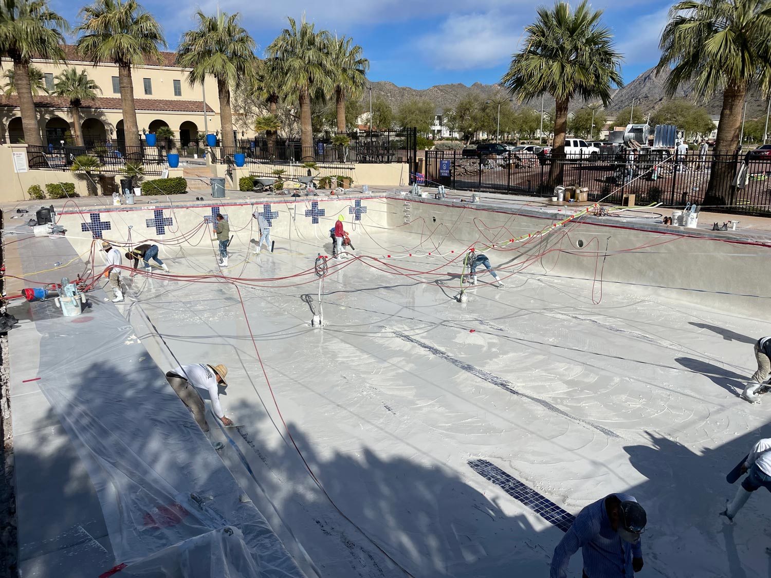 Workers are resurfacing an empty swimming pool, surrounded by palm trees and buildings with mountains in the background.